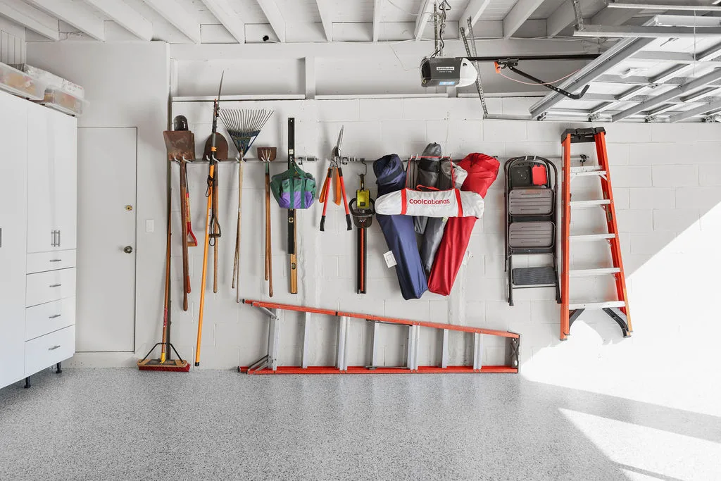 Garage wall organized with custom-mounted tools, yard equipment, and a folding ladder over an epoxy floor